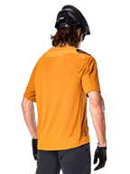 Alpinestars A-Aria Polartec Switch tröja, kortärmad MTB-tröja, Dark Gold, vy bakifrån som visar prestandapassform, bärs med svart hjälm och gråa shorts, slitstarka och ventilerande kläder för stigcykling