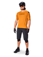 Manlig cyklist i Alpinestars A-Aria Polartec Switch-tröja i Dark Gold, kortärmad MTB-tröja, kombinerad med svarta cykelshorts, knäskydd, svart hjälm och handskar, professionell mountainbike-outfit för stigcykling
