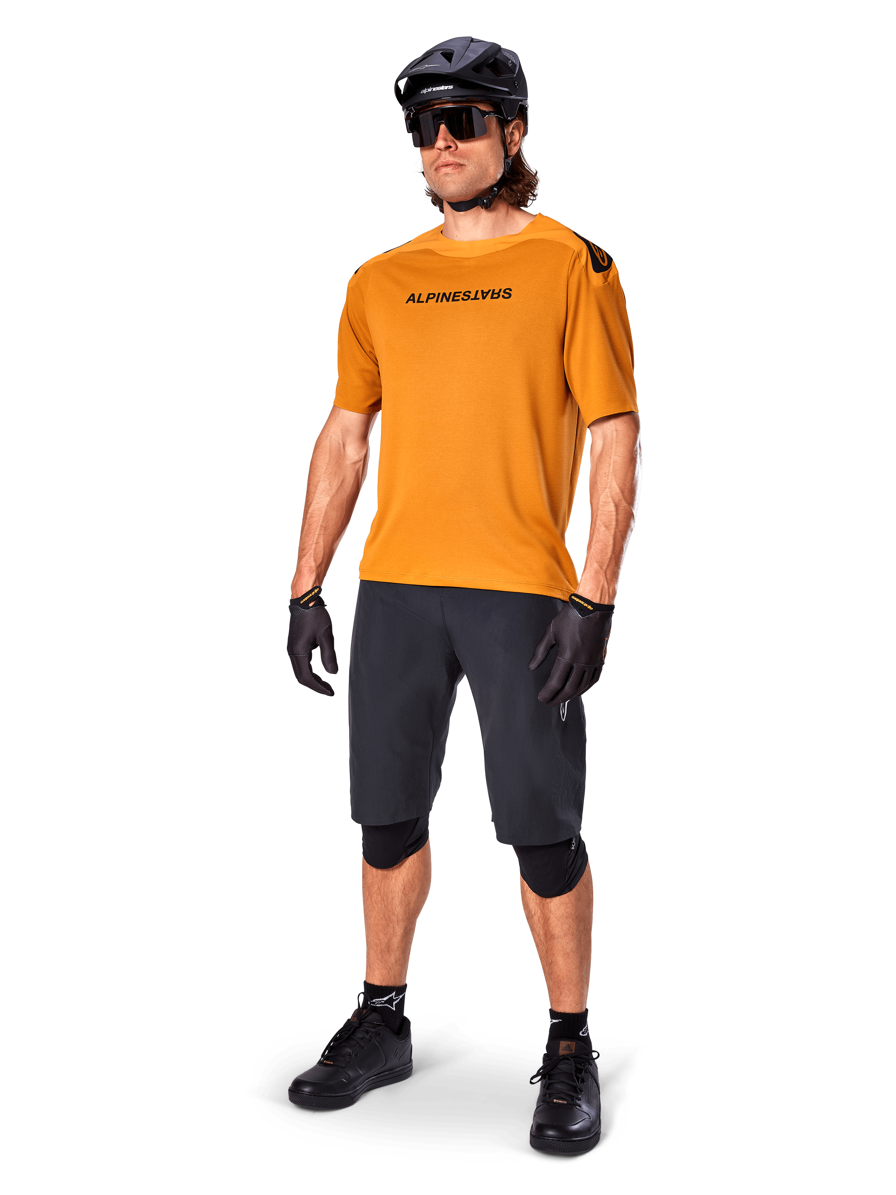 Manlig cyklist i Alpinestars A-Aria Polartec Switch-tröja i Dark Gold, kortärmad MTB-tröja, kombinerad med svarta cykelshorts, knäskydd, svart hjälm och handskar, professionell mountainbike-outfit för stigcykling