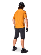 Alpinestars A-Aria Polartec Switch-tröja, kortärmad MTB-tröja, mörkt guld, baksida av modell som bär mörkgrå mountainbike-shorts, svart hjälm och handskar, tekniska cykelkläder för terrängstigar