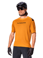 Alpinestars A-Aria Polartec Switch Jersey, kortärmad MTB-tröja, Dark Gold med svart logotyp, bärs av modell med svart hjälm, solglasögon och cykelhandskar, högpresterande mountainbike-kläder
