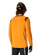 Alpinestars A-Dura Dri Wool långärmad tröja i Dark Gold, sedd bakifrån på en cyklist med mountainbikehjälm och handskar, svarta meshpaneler på axlarna och ett längre ryggslut, tekniska cykelkläder för trailcykling