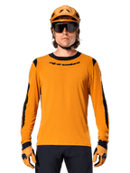 Manlig modell bär Alpinestars A-Dura Dri Wool Long Sleeve Jersey i Dark Gold, matchad med en matchande guld-MTB-hjälm och handskar, svarta meshpaneler på axlar och ärmar, svart Alpinestars-logotyp på bröstet, mörka solglasögon och byxor, tekniska mountainbikekläder