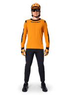 Mountainbike-cyklist bär Alpinestars A-Dura Dri Wool Long Sleeve Jersey, mörkguld med svarta detaljer på krage och ärmar, kombinerat med svarta byxor, matchande guldfärgad hjälm och handskar samt svarta solglasögon, professionell mountainbike-utrustning