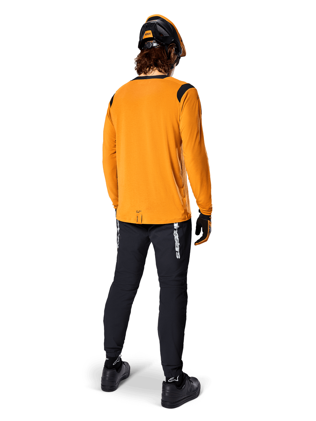 Mountainbikecyklist sedd bakifrån bär Alpinestars A-Dura Dri Wool långärmad tröja i Dark Gold, svarta tekniska byxor med vit logotyp och en matchande svart och orange hjälm, högpresterande mountainbike-kläder.