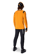 Mountainbikecyklist sedd bakifrån bär Alpinestars A-Dura Dri Wool långärmad tröja i Dark Gold, svarta tekniska byxor med vit logotyp och en matchande svart och orange hjälm, högpresterande mountainbike-kläder.