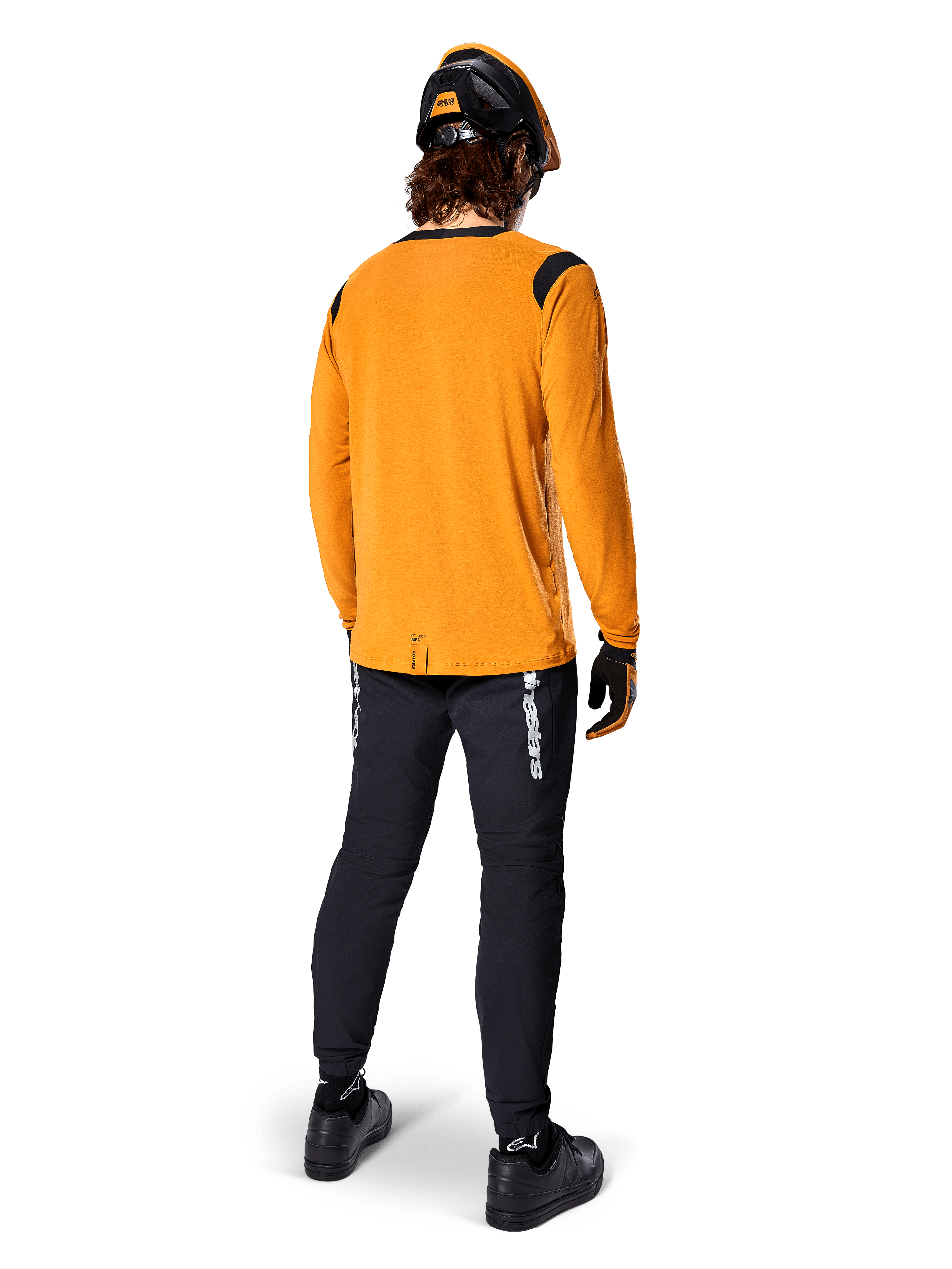Mountainbikecyklist sedd bakifrån bär Alpinestars A-Dura Dri Wool långärmad tröja i Dark Gold, svarta tekniska byxor med vit logotyp och en matchande svart och orange hjälm, högpresterande mountainbike-kläder.