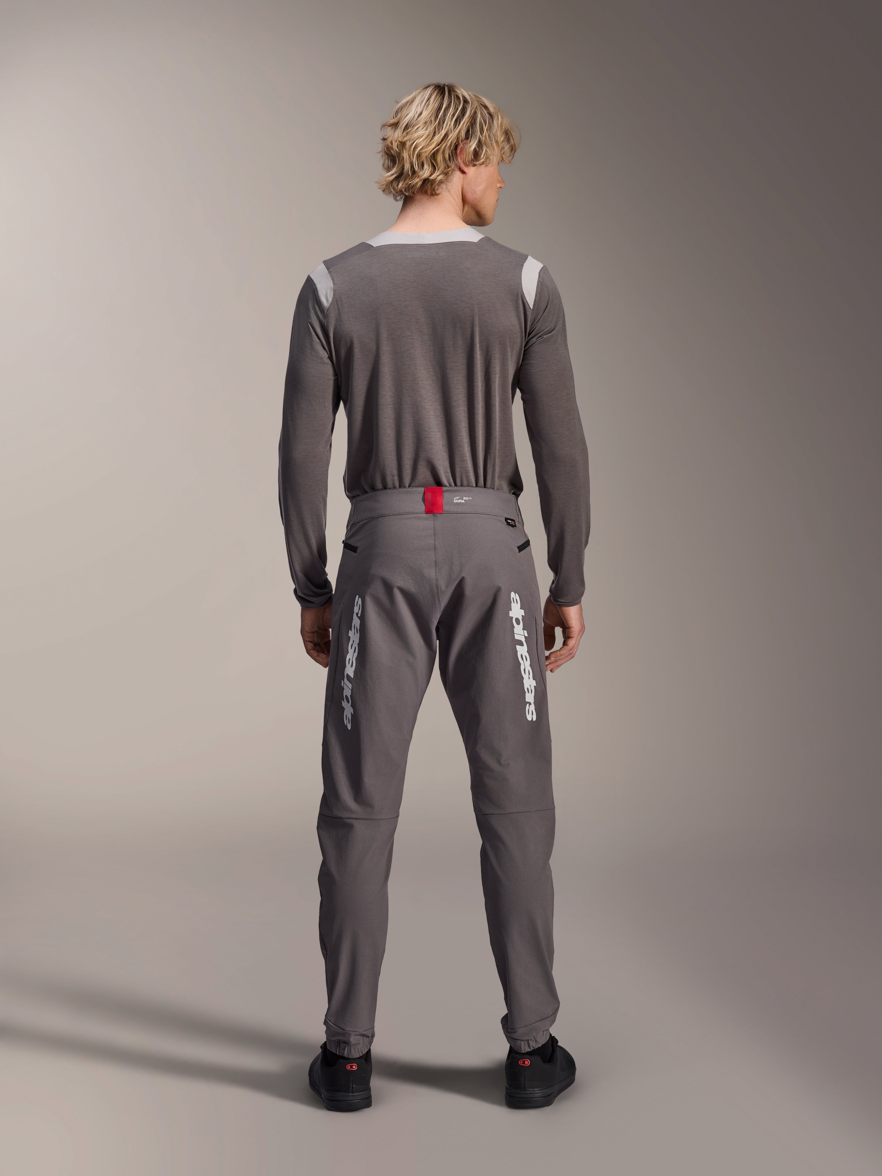 Model from behind wearing Alpinestars A-Dura Elite Pants in Dark Gray and a matching gray long-sleeve jersey, featuring white Alpinestars logos on the legs, a red accent on the waistband, and black shoes, professional MTB riding gear.