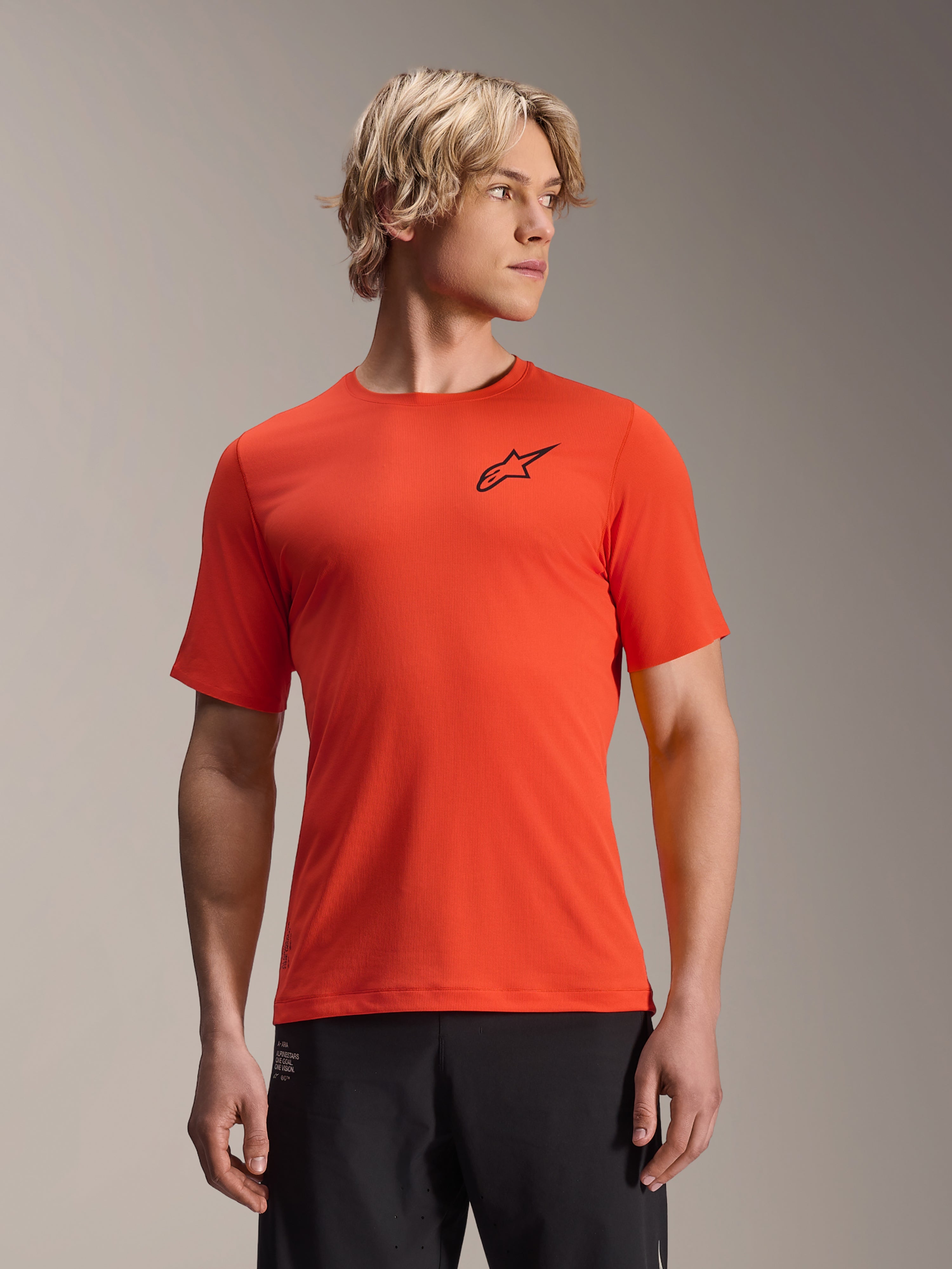 Alpinestars A-Aria Astar kortärmad tröja, Spicy Orange med svart stjärnlogotyp, MTB-tröja för herrar, prestandapassform, bärs av en manlig modell med blont hår och svarta byxor