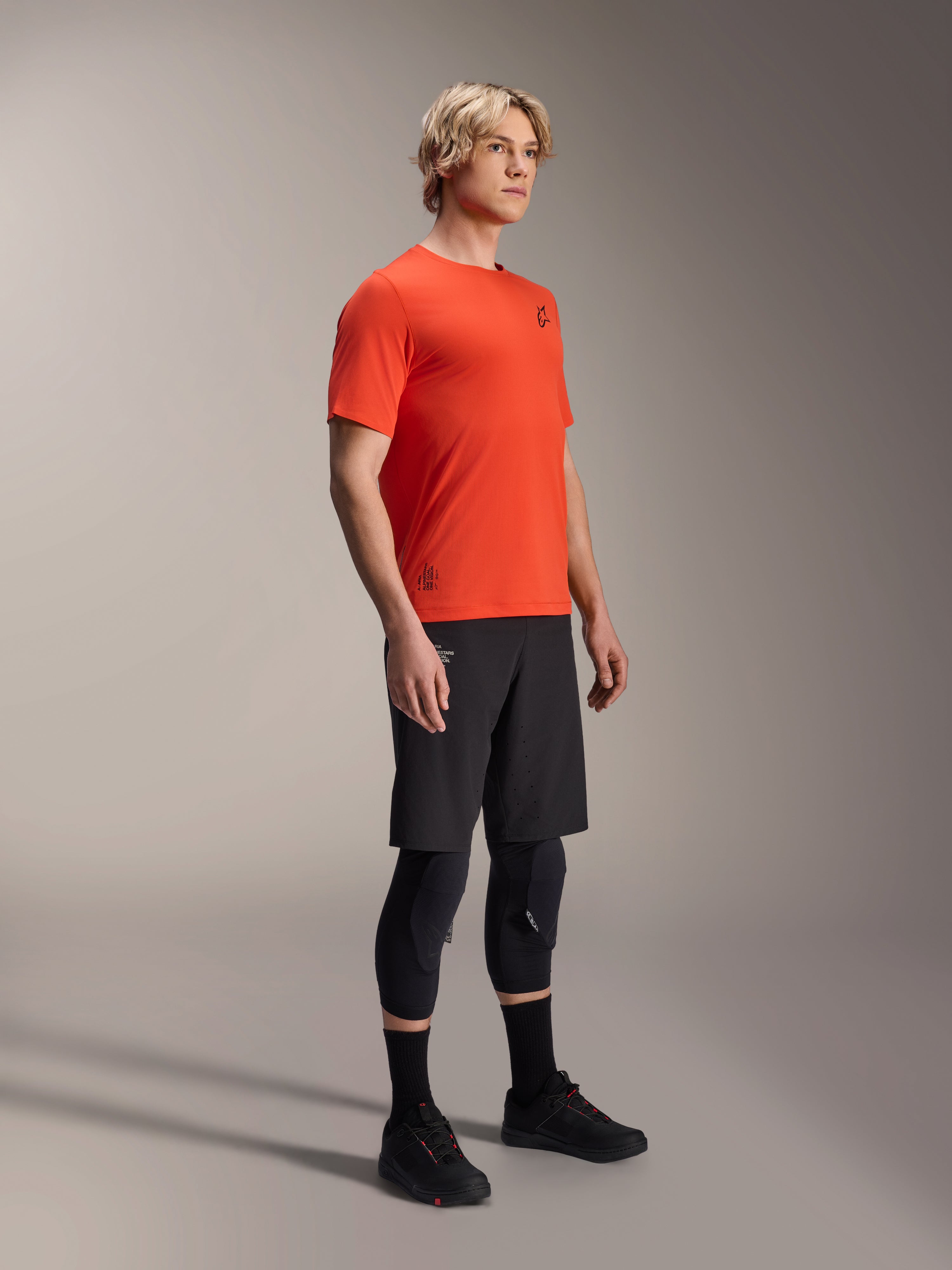 Alpinestars A-Aria Astar kortärmad tröja i Spicy Orange, bärs av en modell med svarta MTB-shorts och knäskydd, lätta och ventilerande mountainbike-kläder, livfull orange med minimal svart logotyp