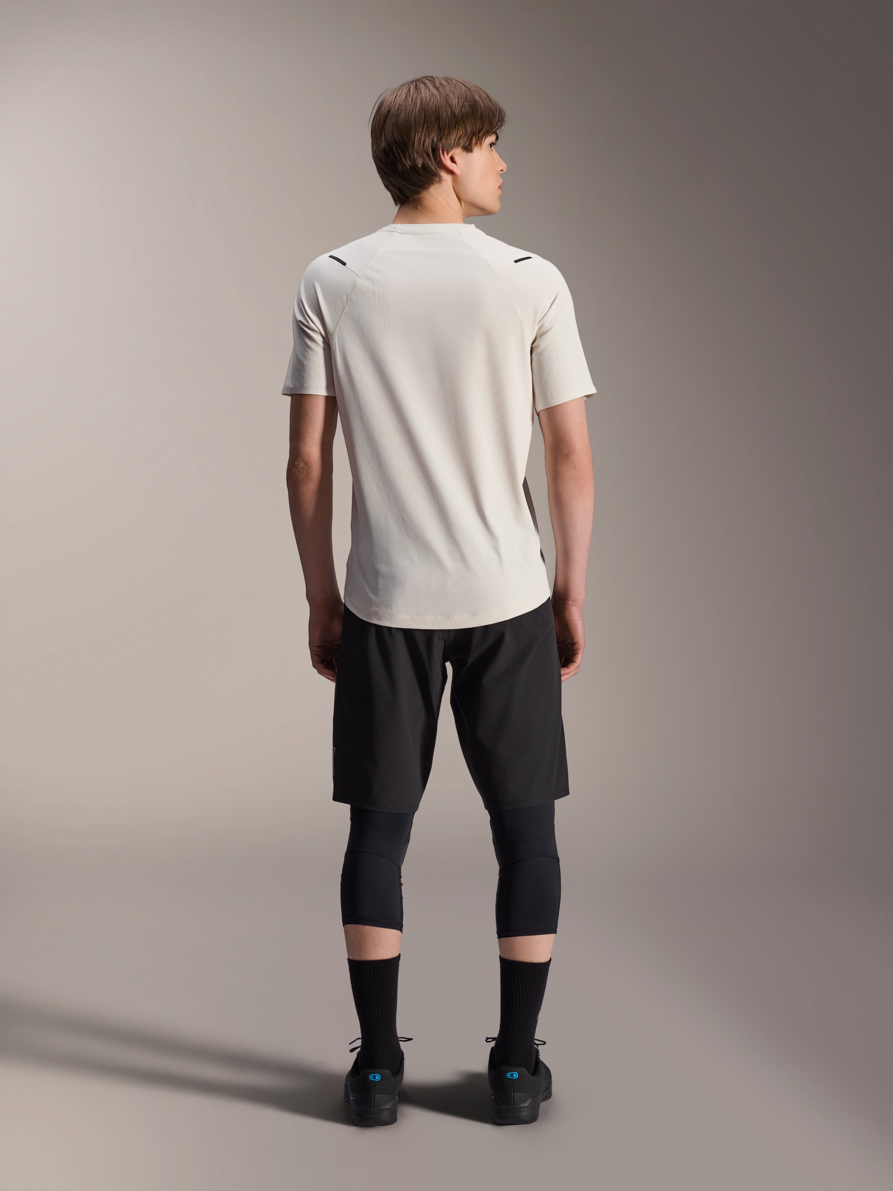 Manlig modell sedd bakifrån bärande Alpinestars A-Aria Astar kortärmad tröja i Ecru, kombinerad med svarta MTB-shorts och skyddande leggings, med förlängd rygg och prestandapassform för stigcykling.