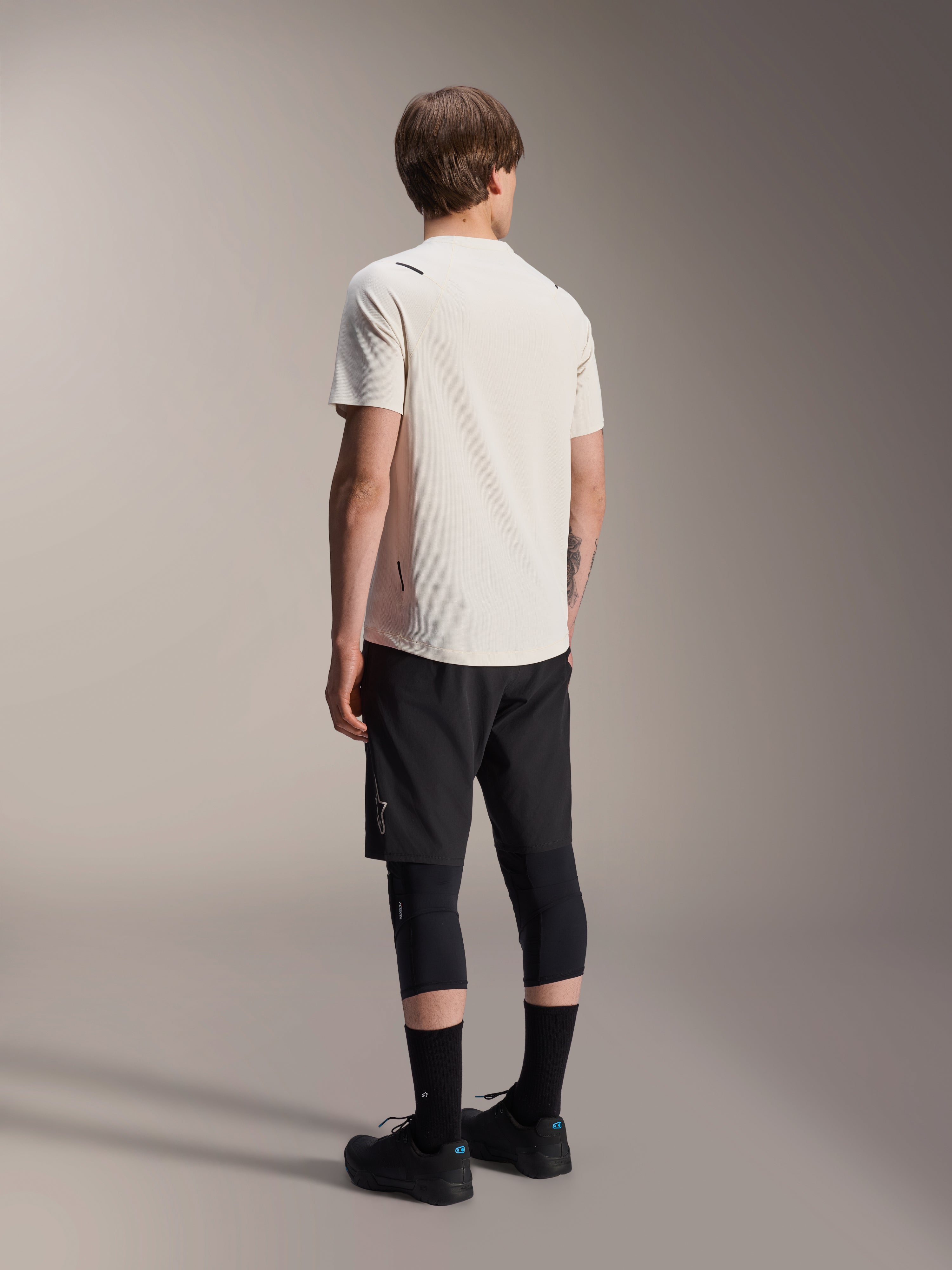 Alpinestars A-Aria Astar Jersey, kortärmad MTB-tröja, Ecru, vy bakifrån som visar prestandapassform och förlängd rygg, stylad med svarta mountainbike-shorts, knäskydd och svarta cykelskor för stigcykling