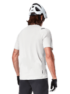 Alpinestars A-Aria Astar Jersey, kortärmad, White Dove, vy bakifrån av en lätt och andningsbar mountainbike-tröja, bärs med en vit hjälm och svarta cykelhandskar, med ett förlängt bakstycke för extra täckning