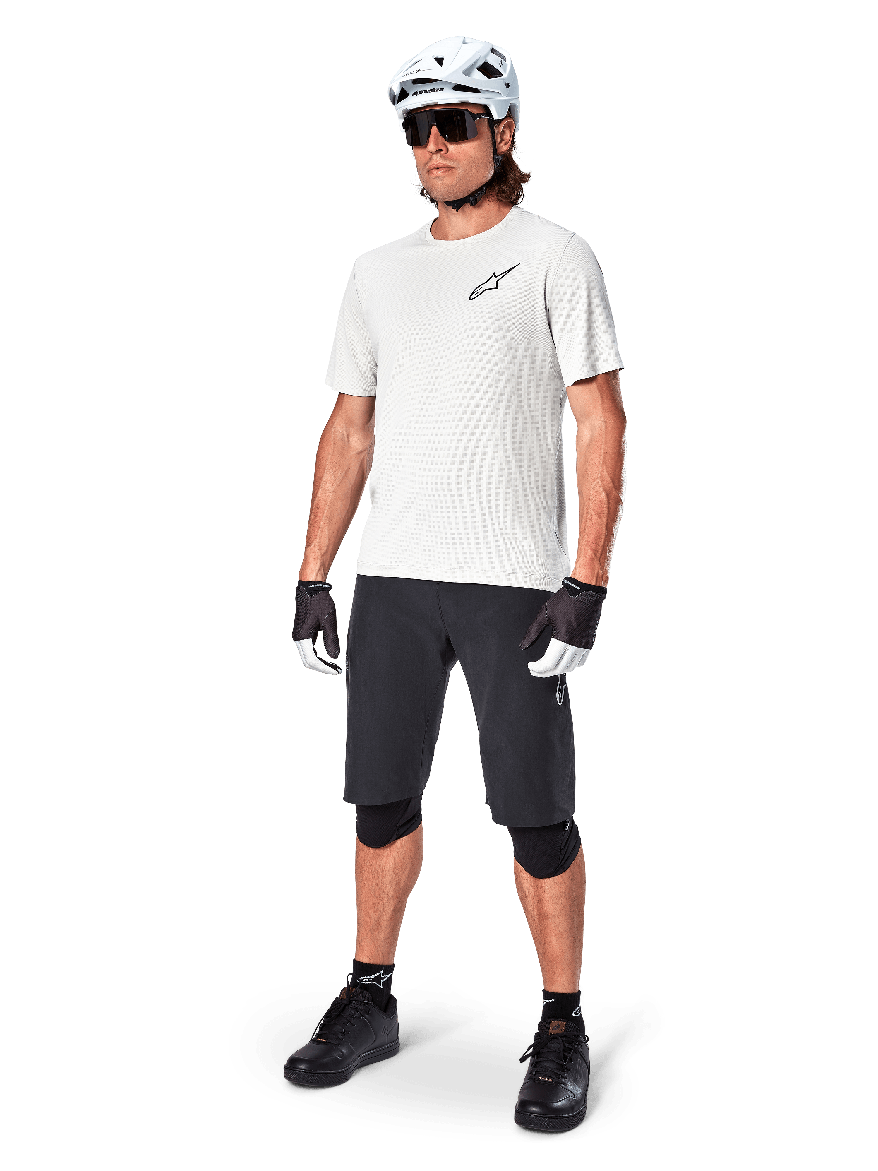 MTB-cyklist iklädd Alpinestars A-Aria Astar kortärmad tröja i White Dove, kombinerad med svarta mountainbikeshorts, vit hjälm och svarta knäskydd, med en liten svart logotyp på bröstet, matchande cykelutrustning för stigcykling