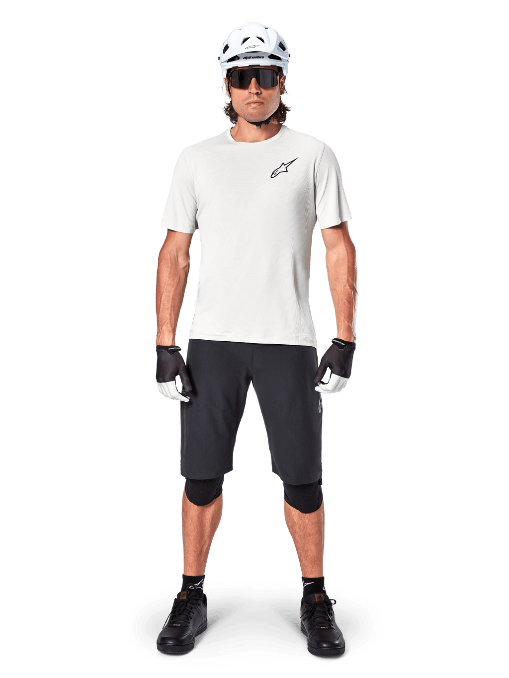 Manlig cyklist som bär Alpinestars A-Aria Astar kortärmad tröja i White Dove, med en svart Astars-logotyp på bröstet, matchad med svarta mountainbikeshorts, vit hjälm, solglasögon, svarta knäskydd och handskar, komplett outfit för terrängcykling.