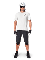 Manlig cyklist som bär Alpinestars A-Aria Astar kortärmad tröja i White Dove, med en svart Astars-logotyp på bröstet, matchad med svarta mountainbikeshorts, vit hjälm, solglasögon, svarta knäskydd och handskar, komplett outfit för terrängcykling.