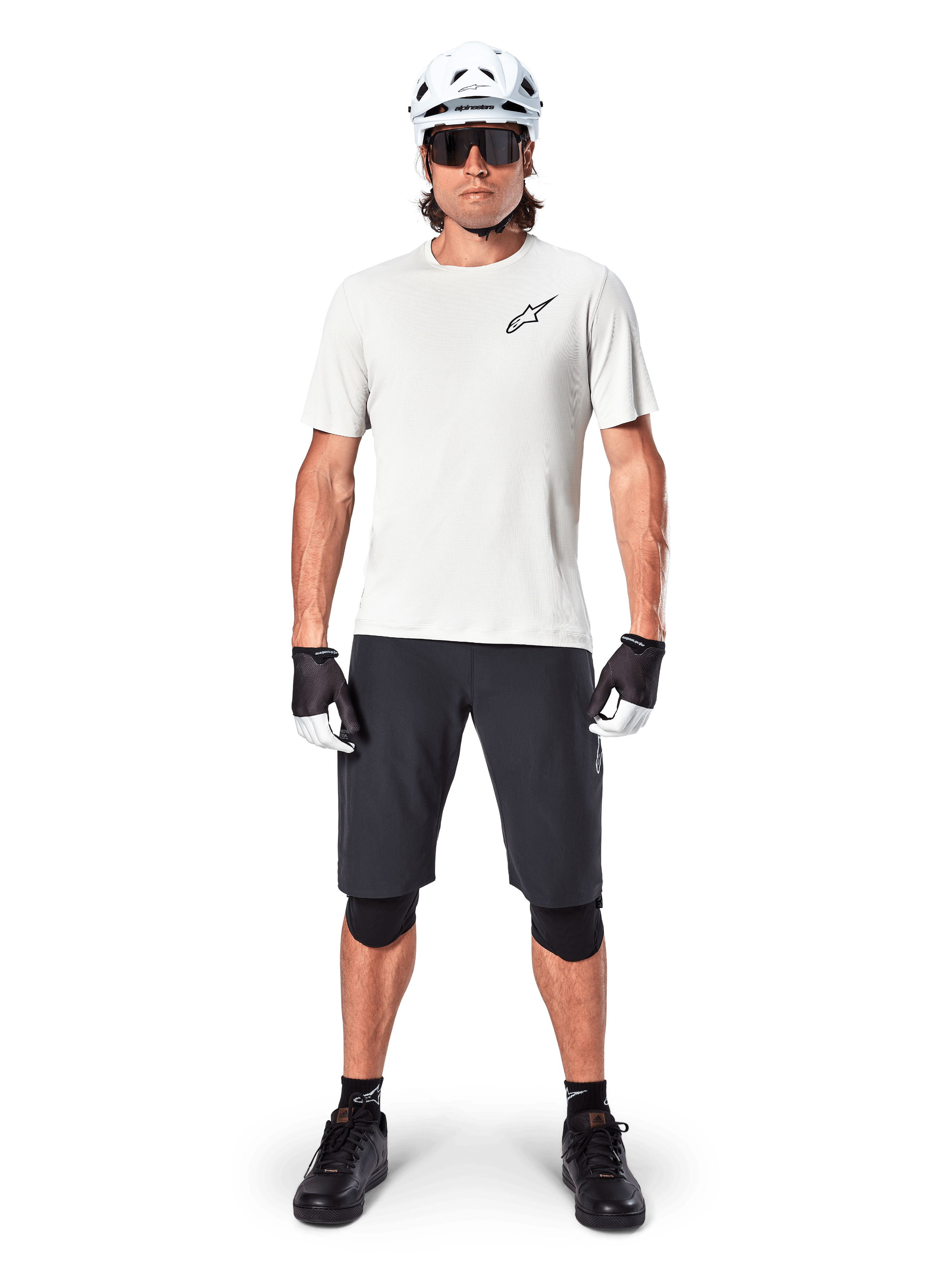Manlig cyklist som bär Alpinestars A-Aria Astar kortärmad tröja i White Dove, med en svart Astars-logotyp på bröstet, matchad med svarta mountainbikeshorts, vit hjälm, solglasögon, svarta knäskydd och handskar, komplett outfit för terrängcykling.
