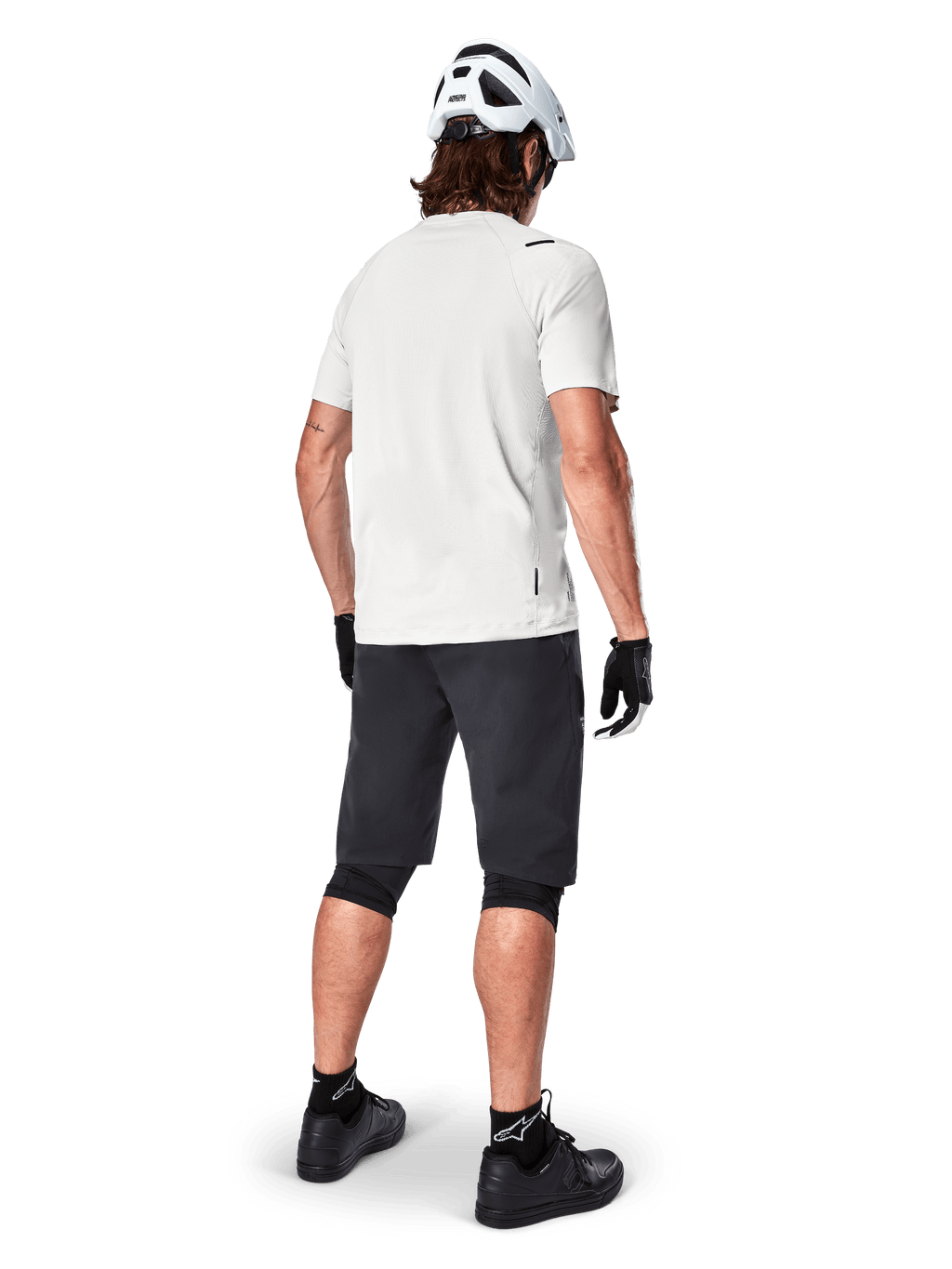 Alpinestars A-Aria Astar kortärmad tröja i White Dove, cyklist sedd bakifrån, kombinerat med svarta MTB-shorts, knäskydd och en vit hjälm, lätt och ventilerande utrustning för terrängcykling