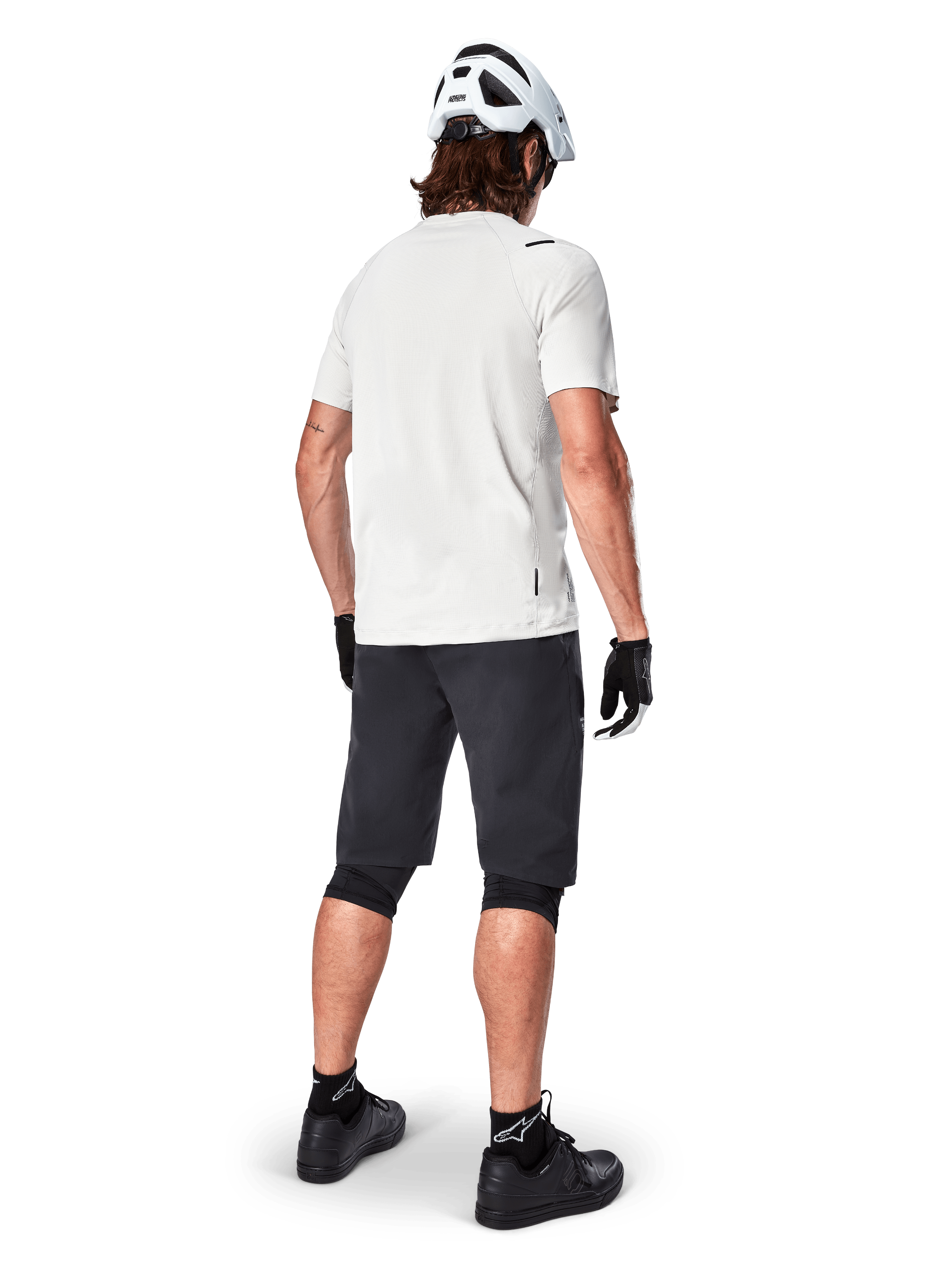 Alpinestars A-Aria Astar kortärmad tröja i White Dove, cyklist sedd bakifrån, kombinerat med svarta MTB-shorts, knäskydd och en vit hjälm, lätt och ventilerande utrustning för terrängcykling