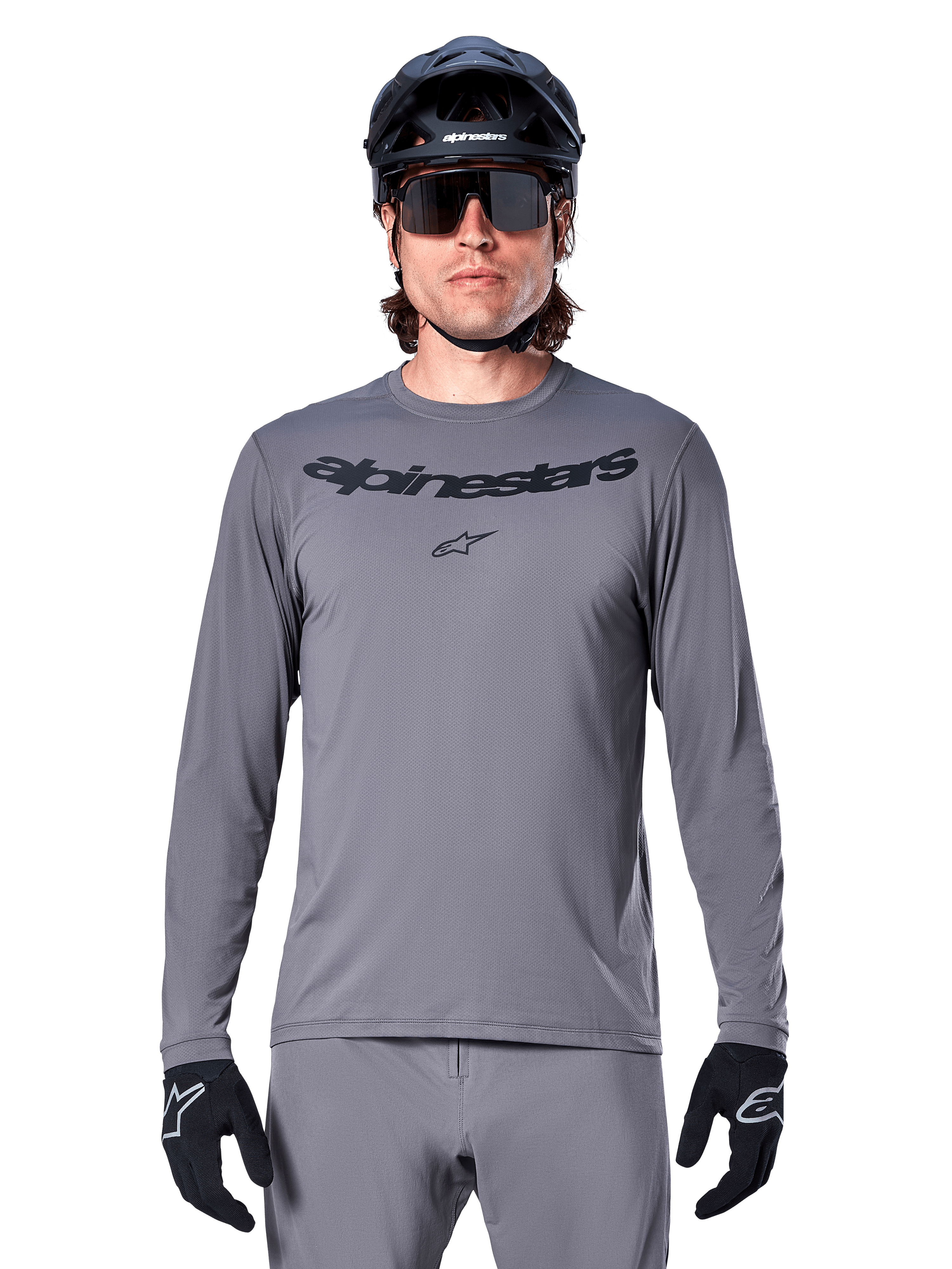 Male rider wearing the Alpinestars A-Dura Rocker Long Sleeve Jersey, Dark Gray MTB jersey with black wordmark on chest, paired with gray pants, black helmet, and gloves, designed for off-road cycling