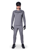 Male cyclist wearing Alpinestars A-Dura Rocker Long Sleeve Jersey in Dark Gray, matching gray pants, black MTB helmet, sunglasses and gloves, professional mountain bike apparel for off-road trails
