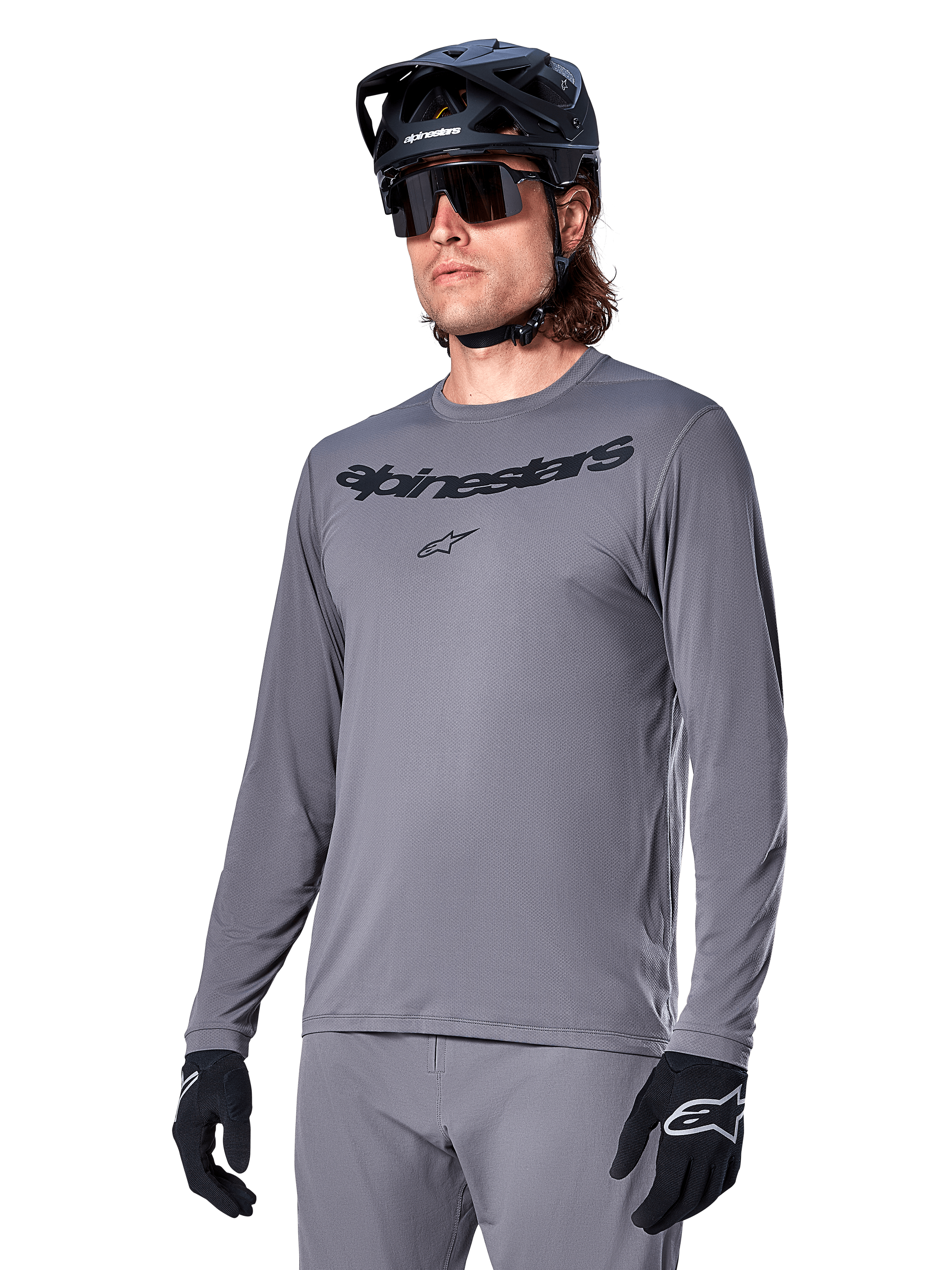Alpinestars A-Dura Rocker långärmad tröja, grå MTB-tröja, bärs med svart hjälm och handskar, med svart ordmärke på bröstet, sportiga cykelkläder för terrängkörning, enfärgad bakgrund