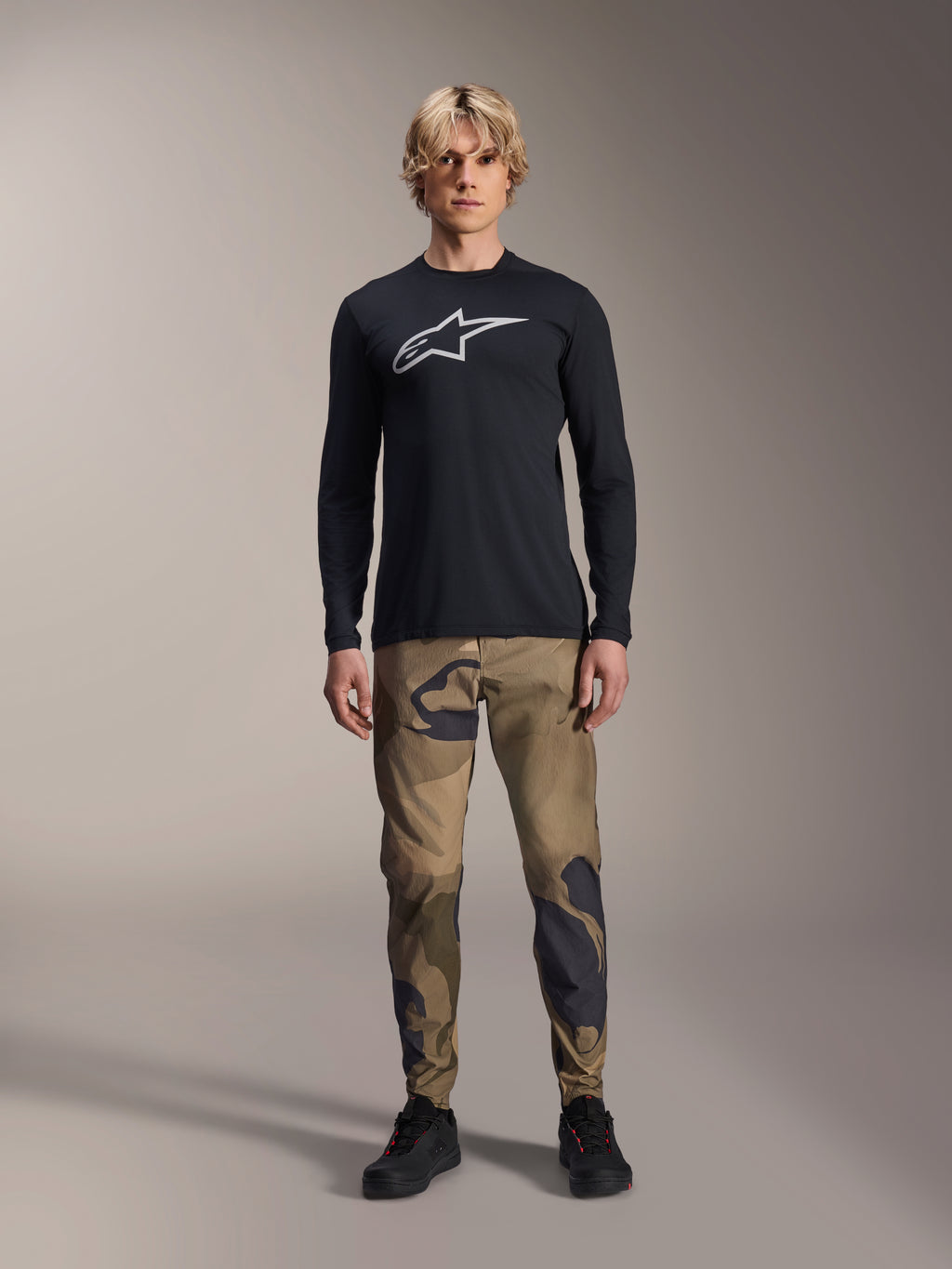 Alpinestars A-Dura Astar långärmad tröja, svart, med en stor vit Astars-logotyp på bröstet, bärs med kamouflagebyxor och svarta skor, avslappnade funktionskläder för mountainbike