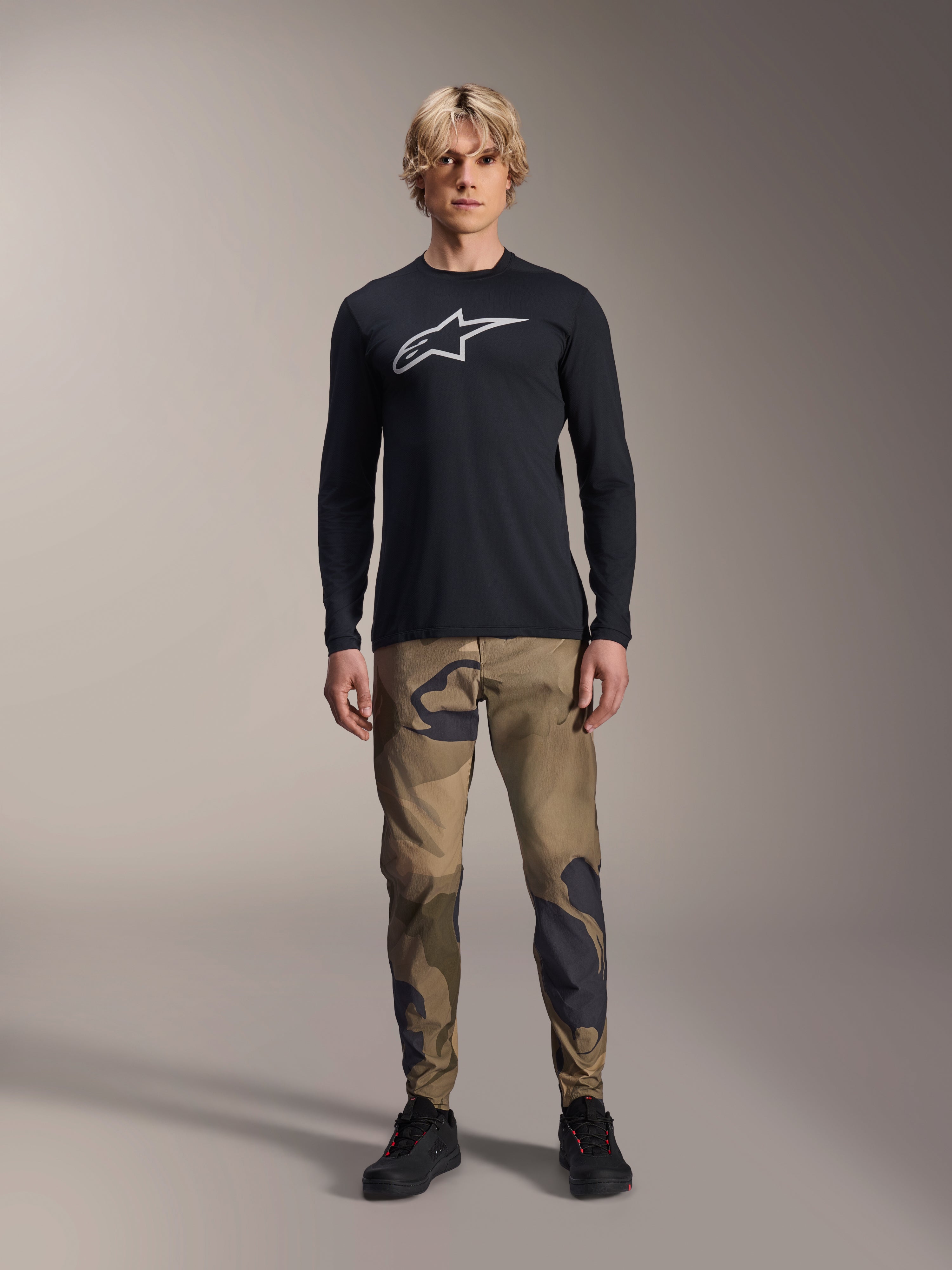 Alpinestars A-Dura Astar långärmad tröja, svart, med en stor vit Astars-logotyp på bröstet, bärs med kamouflagebyxor och svarta skor, avslappnade funktionskläder för mountainbike
