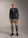 Alpinestars A-Dura Camo Pant, MTB-cykelbyxor, brunt och grönt kamouflage, bärs med en svart långärmad tröja och svarta skor, sportig mountainbikestil med slitstarkt stretchmaterial