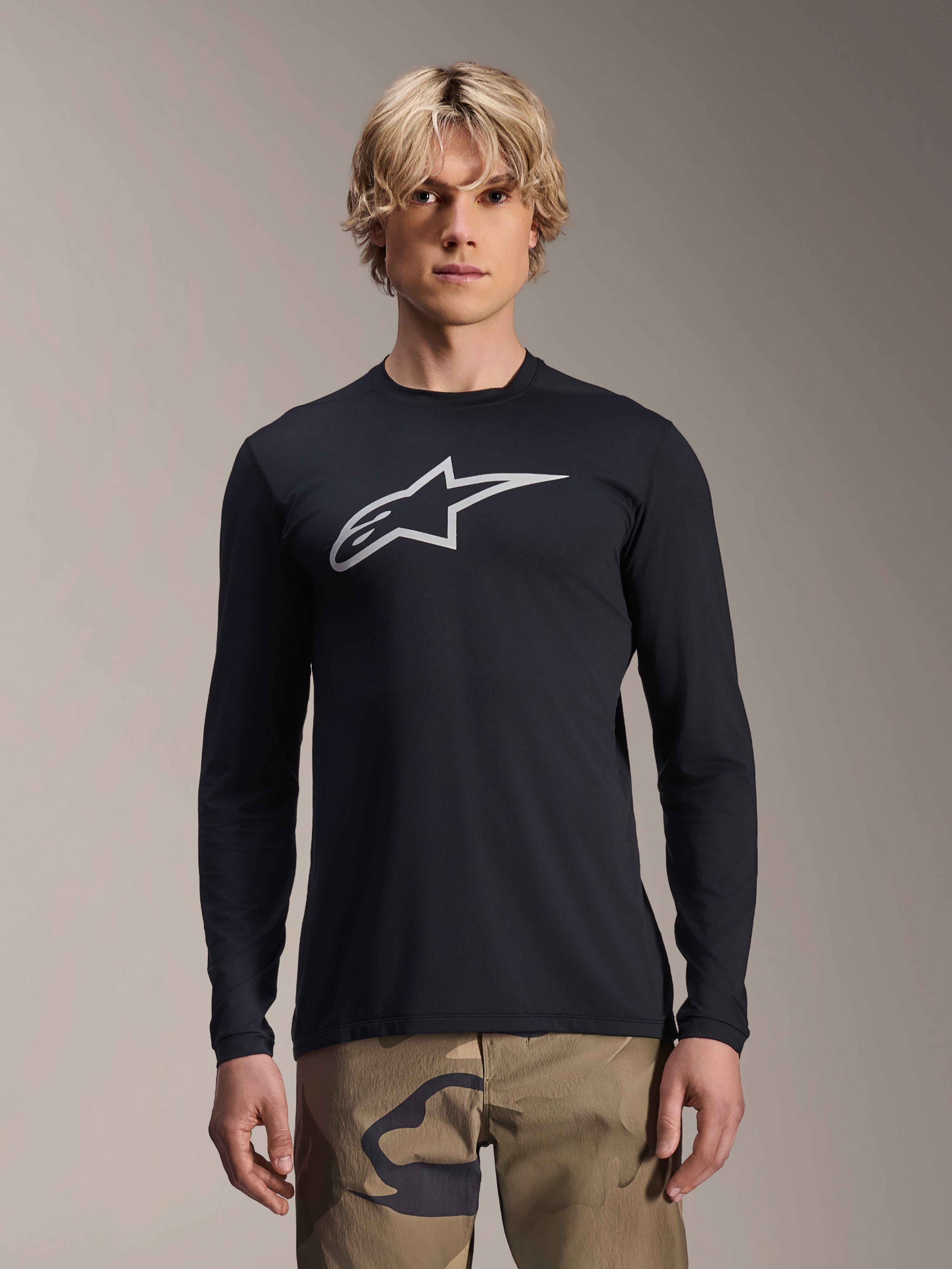 Alpinestars A-Dura Astar Long Sleeve Jersey, Black with silver Astars logo on the chest, worn by a model with camouflage pants, casual MTB performance apparel for mountain biking