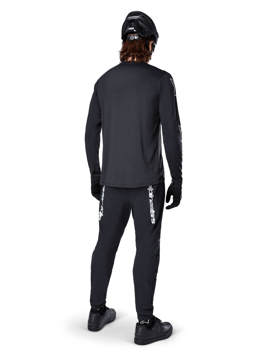 Alpinestars A-Dura Switch långärmad tröja, svart, vy bakifrån på en cyklist, bärs med svarta byxor med vit Alpinestars-logotyp, svart hjälm och handskar, med förlängt bakstycke för extra täckning, prestandakläder för mountainbike