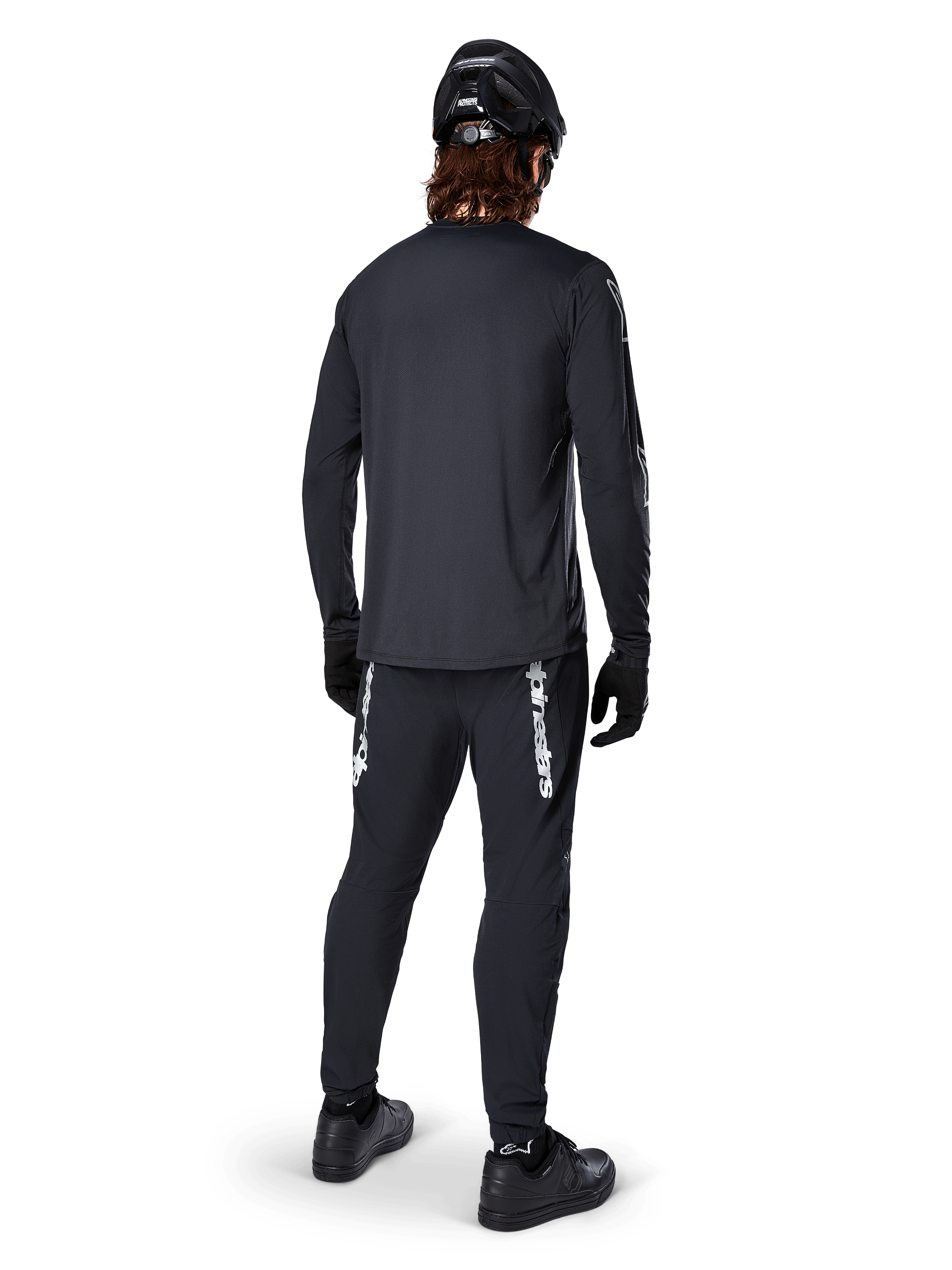 Alpinestars A-Dura Switch långärmad tröja, svart, vy bakifrån på en cyklist, bärs med svarta byxor med vit Alpinestars-logotyp, svart hjälm och handskar, med förlängt bakstycke för extra täckning, prestandakläder för mountainbike