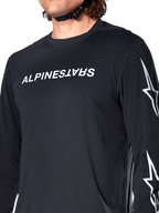 Alpinestars A-Dura Switch långärmad tröja, svart, med vit Alpinestars-ordmärkeslogotyp på bröstet och stjärnlogotyper på ärmen, buren av en cyklist med synlig hjälmrem, tekniska mountainbike-kläder