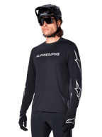 Manlig cyklist i Alpinestars A-Dura Switch långärmad tröja, svart med vit ordmärkeslogotyp och stjärnlogotyper på ärmarna, kombinerad med svart MTB-hjälm och solglasögon, högpresterande cykelutrustning för terrängcykling