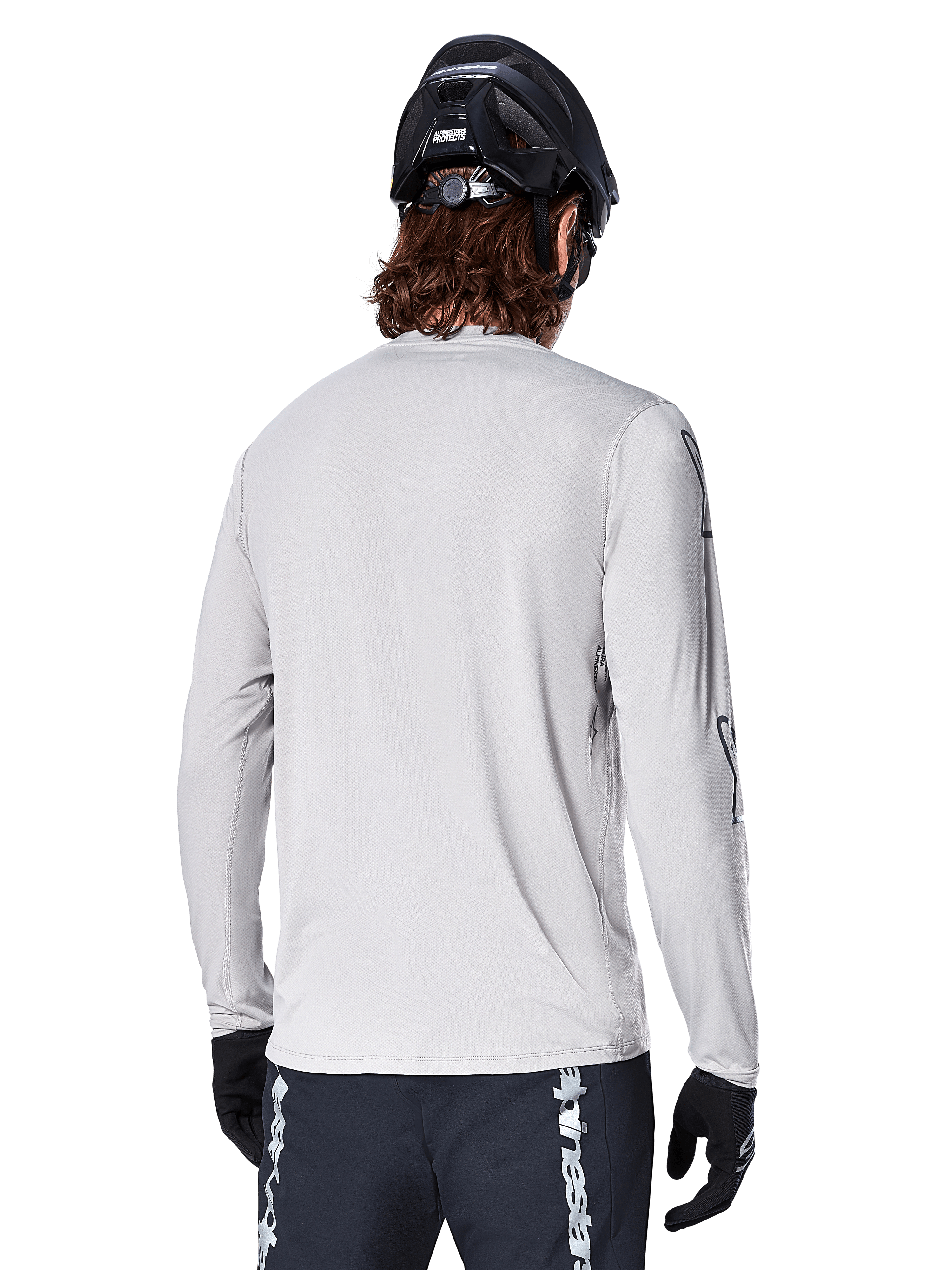 Alpinestars A-Dura Switch långärmad tröja, ljusgrå, bakifrån sett på en mountainbikecyklist med svart MTB-hjälm och handskar, har ett förlängt bakstycke för extra skydd och strukturerat jacquardtyg, teknisk cykelutrustning.