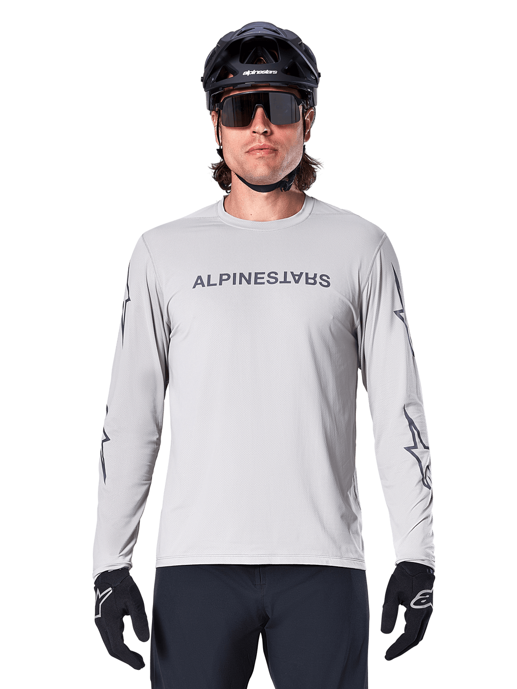 Alpinestars A-Dura Switch långärmad tröja, ljusgrå, mörkgrått ordmärke på bröstet och stjärnlogotyper på ärmarna, bärs av en cyklist med svart MTB-hjälm och handskar, funktionella cykelkläder för stigcykling