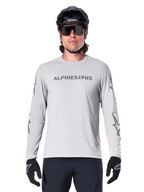 Alpinestars A-Dura Switch långärmad tröja, ljusgrå, mörkgrått ordmärke på bröstet och stjärnlogotyper på ärmarna, bärs av en cyklist med svart MTB-hjälm och handskar, funktionella cykelkläder för stigcykling