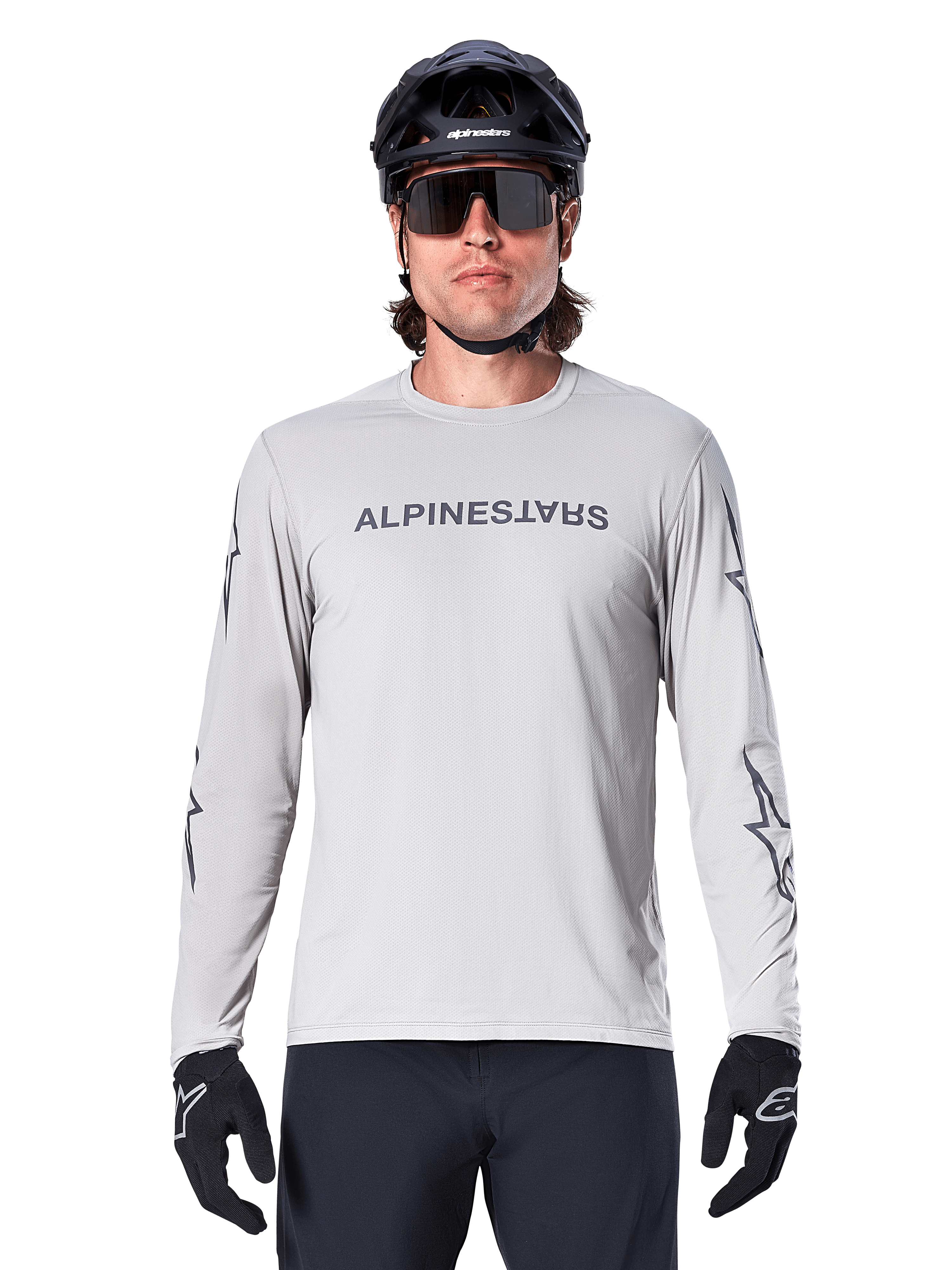 Alpinestars A-Dura Switch långärmad tröja, ljusgrå, mörkgrått ordmärke på bröstet och stjärnlogotyper på ärmarna, bärs av en cyklist med svart MTB-hjälm och handskar, funktionella cykelkläder för stigcykling