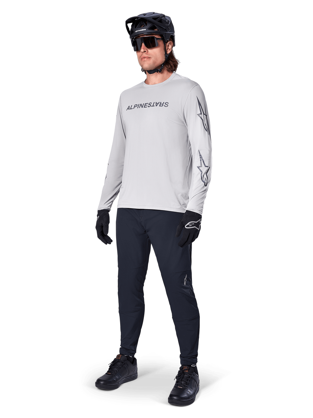 Alpinestars A-Dura Switch långärmad tröja, ljusgrå, bärs av manlig cyklist med svarta mountainbikebyxor och hjälm, med svart logotyp på bröstet och stjärngrafik på ärmarna, högpresterande cykelutrustning för terrängkörning