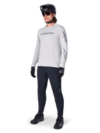 Alpinestars A-Dura Switch långärmad tröja, ljusgrå, bärs av manlig cyklist med svarta mountainbikebyxor och hjälm, med svart logotyp på bröstet och stjärngrafik på ärmarna, högpresterande cykelutrustning för terrängkörning