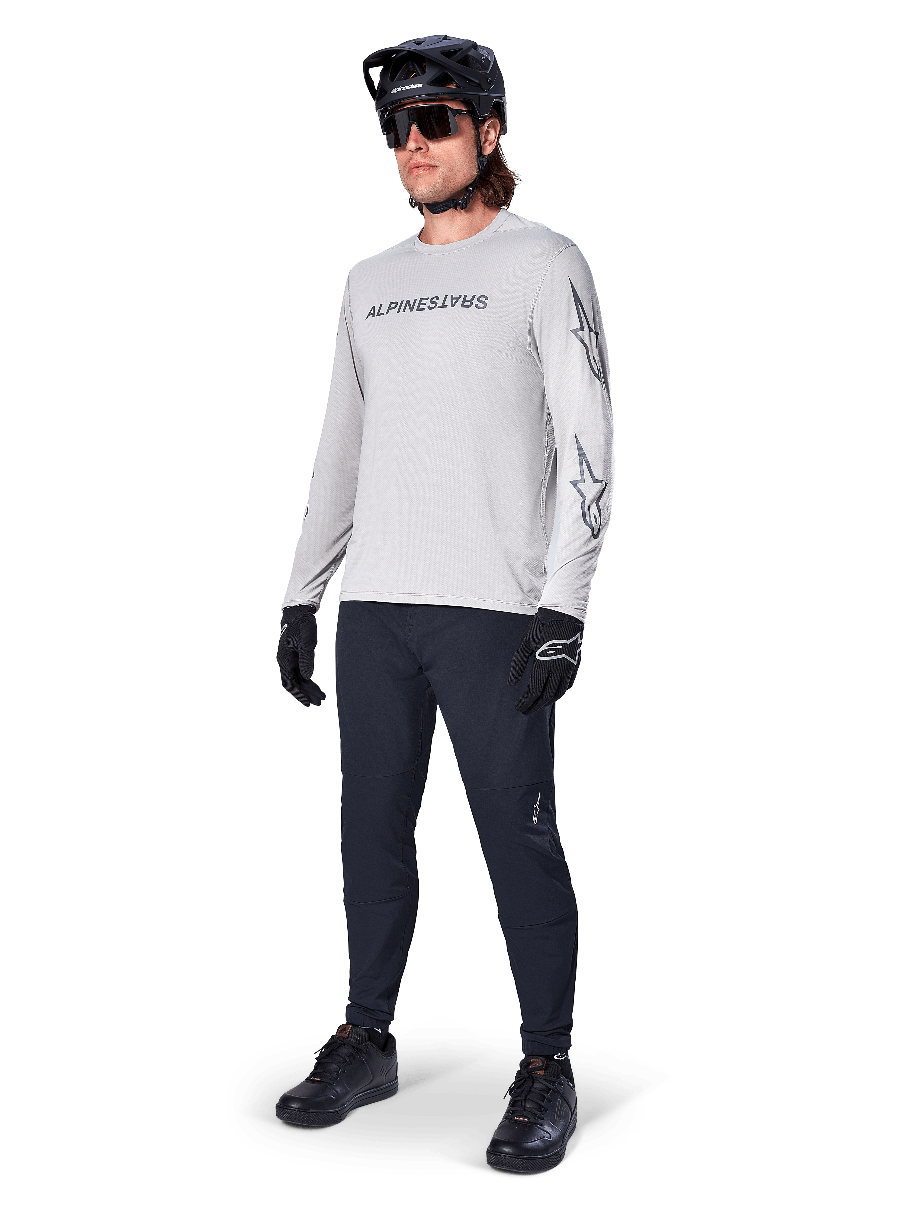 Alpinestars A-Dura Switch långärmad tröja, ljusgrå, bärs av manlig cyklist med svarta mountainbikebyxor och hjälm, med svart logotyp på bröstet och stjärngrafik på ärmarna, högpresterande cykelutrustning för terrängkörning