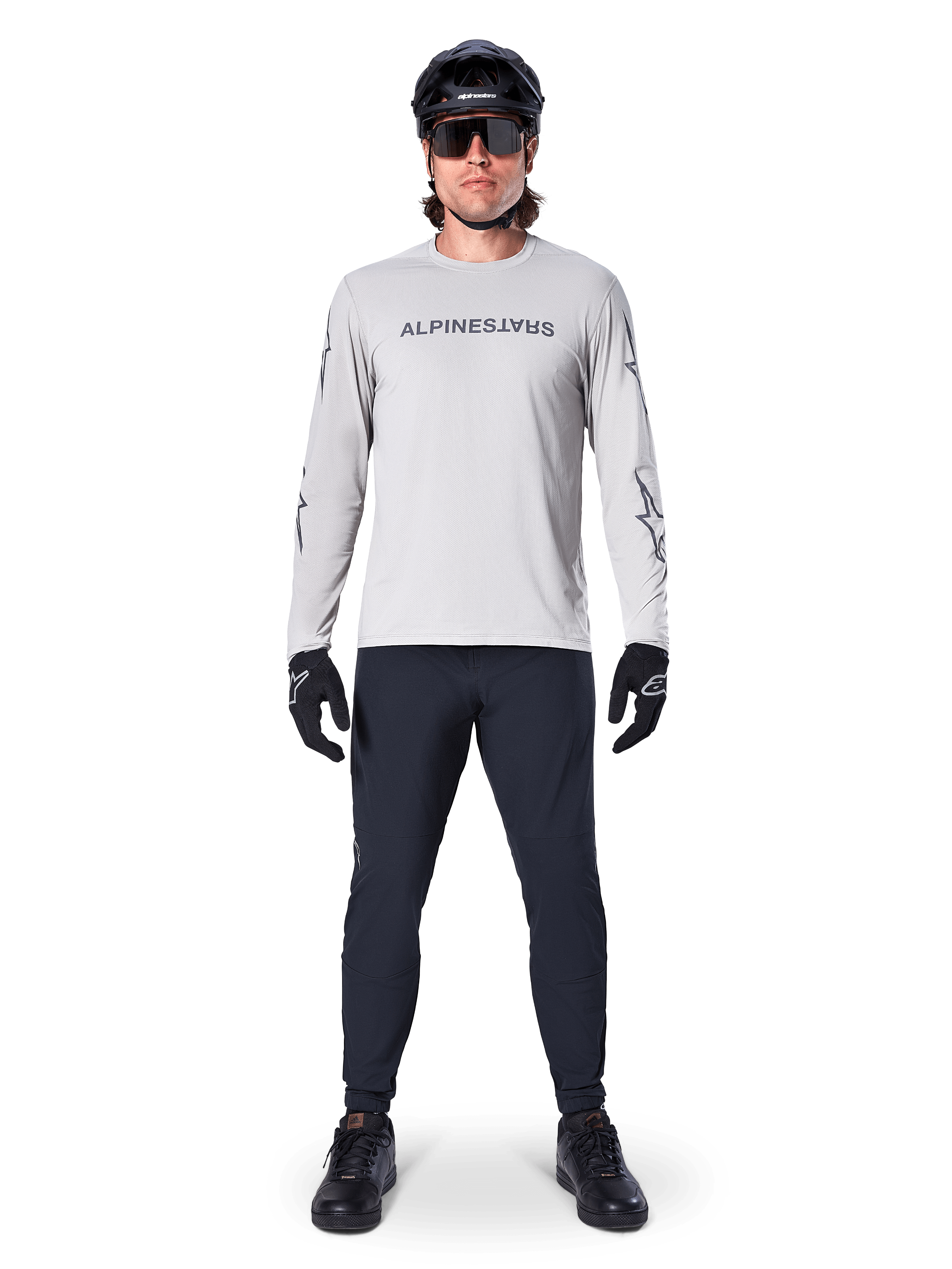 Alpinestars A-Dura Switch långärmad tröja i ljusgrått, med en stiliserad ordmärkeslogotyp på bröstet, bärs med svarta byxor, hjälm och handskar, högpresterande mountainbike-kläder för terrängcykling