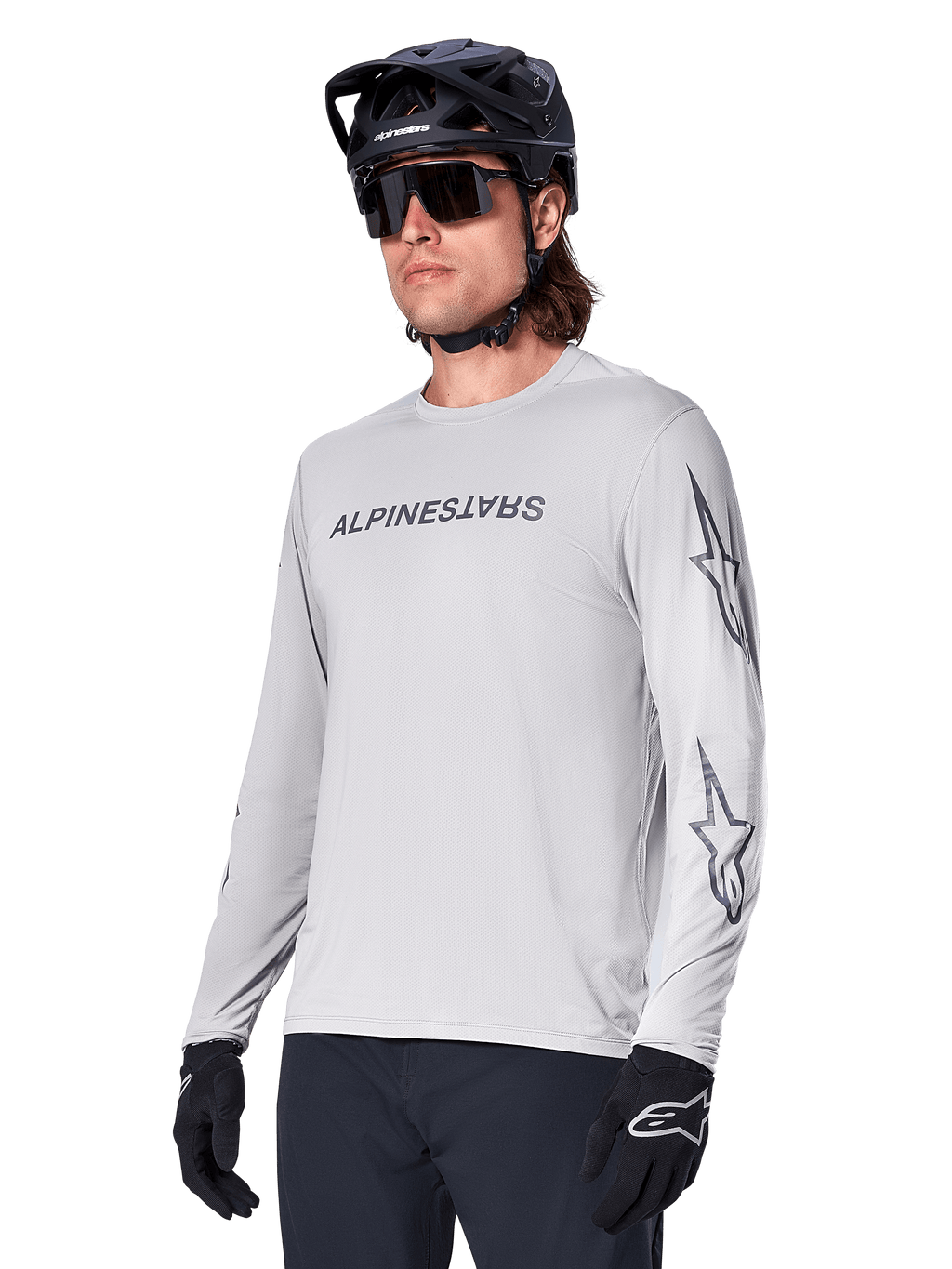 Alpinestars A-Dura Switch långärmad tröja, mountainbiketröja, ljusgrå, svart screentryckt logotyp på bröstet och stjärnlogotyper på ärmarna, bärs med svart mountainbikehjälm, mörka solglasögon och svarta handskar, atletisk cykelutrustning för terrängkörning