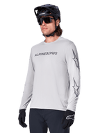 Alpinestars A-Dura Switch långärmad tröja, mountainbiketröja, ljusgrå, svart screentryckt logotyp på bröstet och stjärnlogotyper på ärmarna, bärs med svart mountainbikehjälm, mörka solglasögon och svarta handskar, atletisk cykelutrustning för terrängkörning