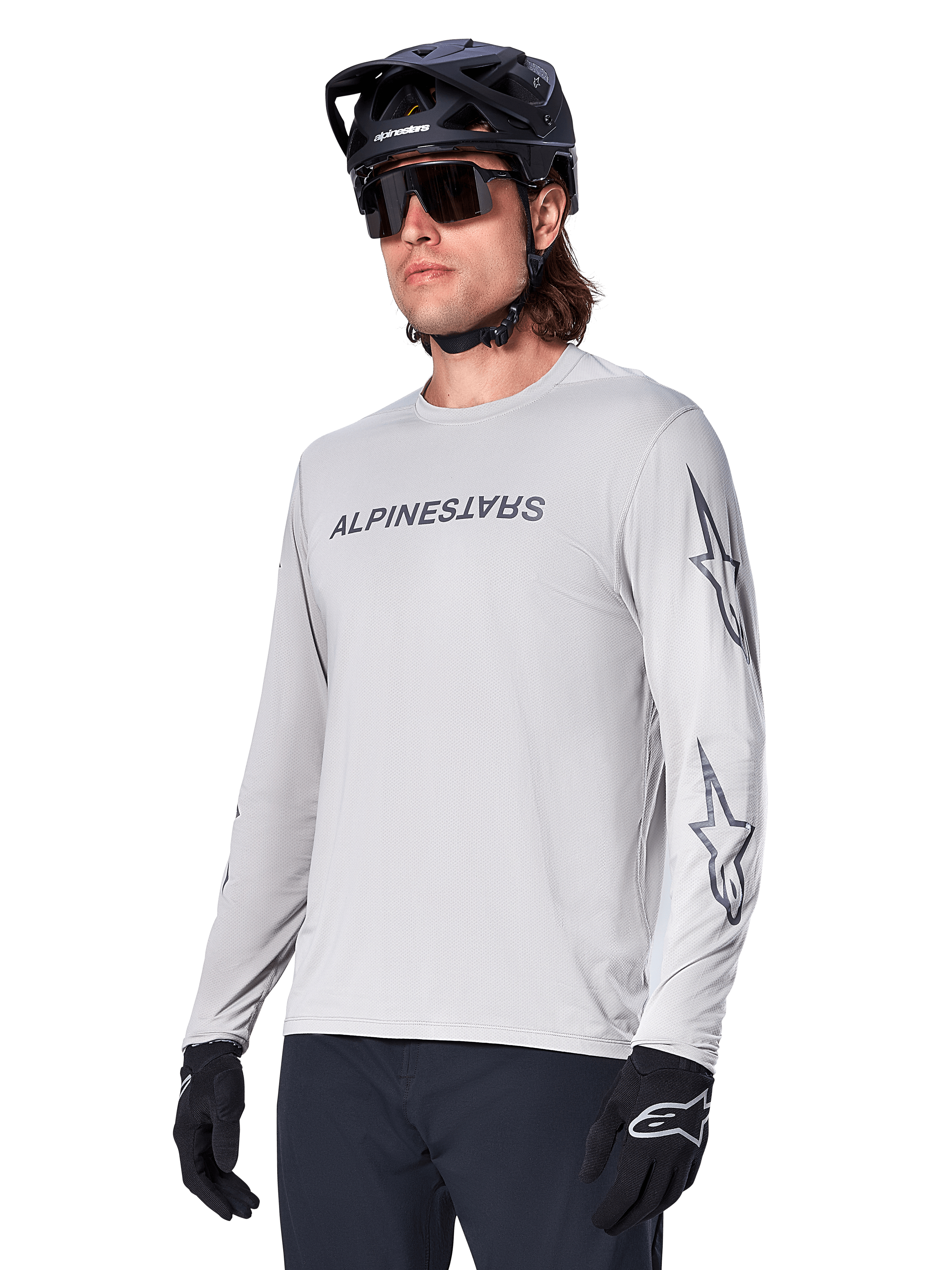 Alpinestars A-Dura Switch långärmad tröja, mountainbiketröja, ljusgrå, svart screentryckt logotyp på bröstet och stjärnlogotyper på ärmarna, bärs med svart mountainbikehjälm, mörka solglasögon och svarta handskar, atletisk cykelutrustning för terrängkörning