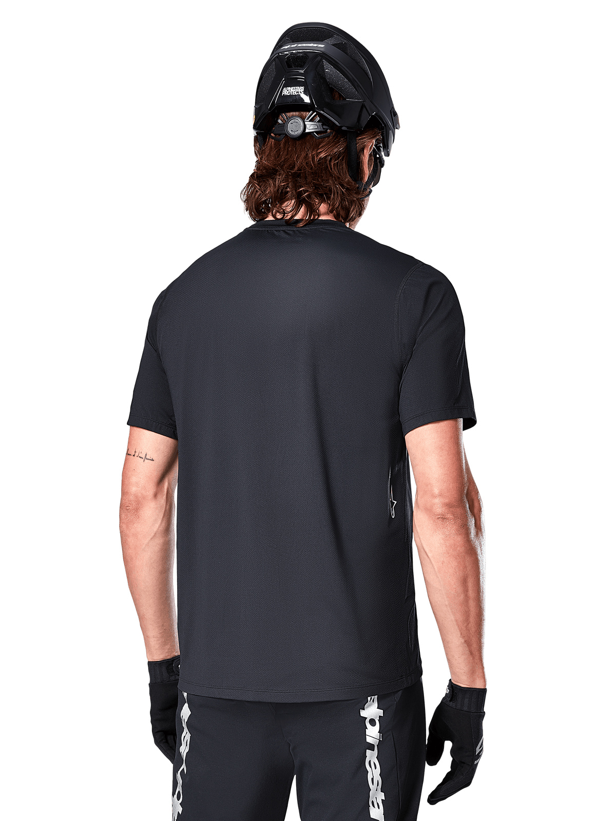 Alpinestars A-Dura Switch kortärmad tröja, svart, vy bakifrån på en modell med svart cykelhjälm och MTB-shorts, framhäver den normala passformen och den förlängda ryggen för mountainbike.