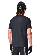 Alpinestars A-Dura Switch kortärmad tröja, svart, vy bakifrån på en modell med svart cykelhjälm och MTB-shorts, framhäver den normala passformen och den förlängda ryggen för mountainbike.