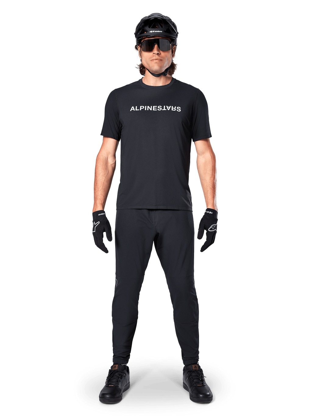 Mountainbike-cyklist i Alpinestars A-Dura Switch kortärmad tröja i svart, med vit stiliserad logotyp på bröstet, matchat med svarta MTB-byxor, hjälm och handskar, komplett cykeloutfit för stigcykling.