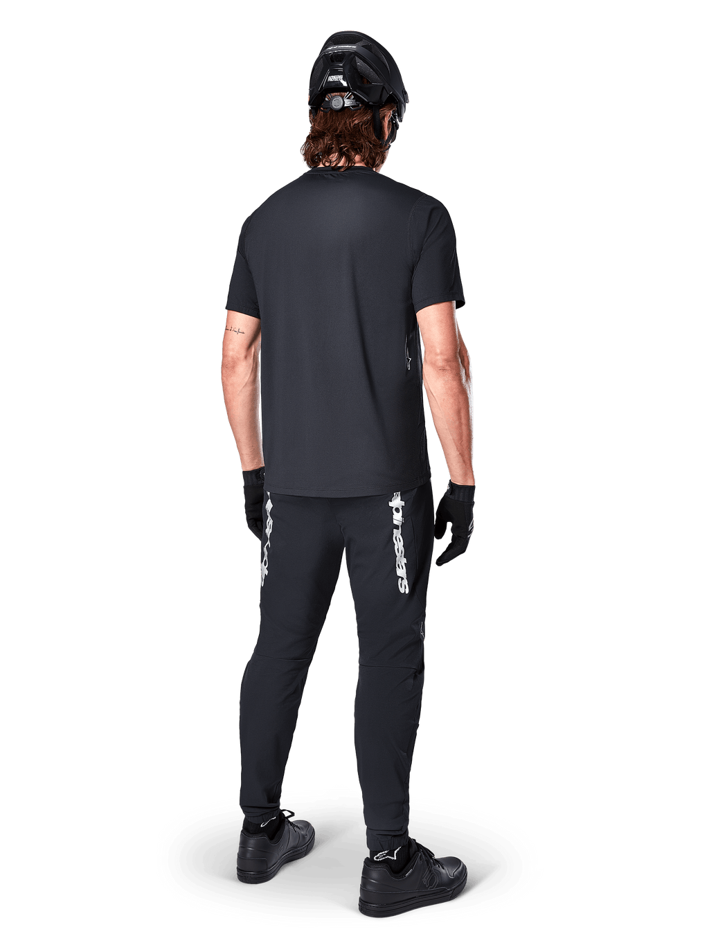 Alpinestars A-Dura Switch-tröja - kortärmad, svart, baksida av cyklist klädd i komplett mountainbike-utrustning, inklusive matchande svarta byxor med vit logotyp, svart hjälm och handskar, tekniska cykelkläder för stigcykling