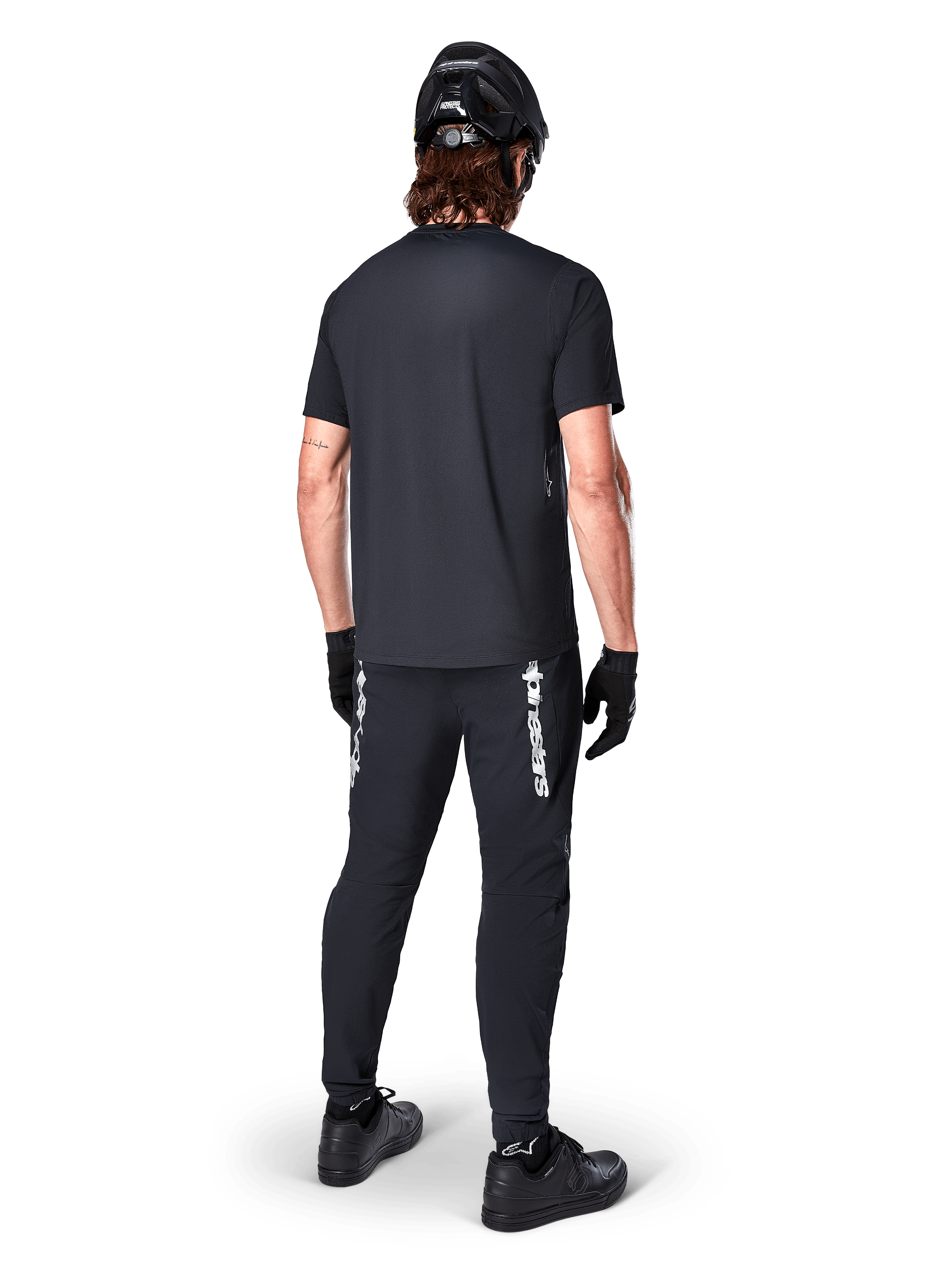 Alpinestars A-Dura Switch-tröja - kortärmad, svart, baksida av cyklist klädd i komplett mountainbike-utrustning, inklusive matchande svarta byxor med vit logotyp, svart hjälm och handskar, tekniska cykelkläder för stigcykling